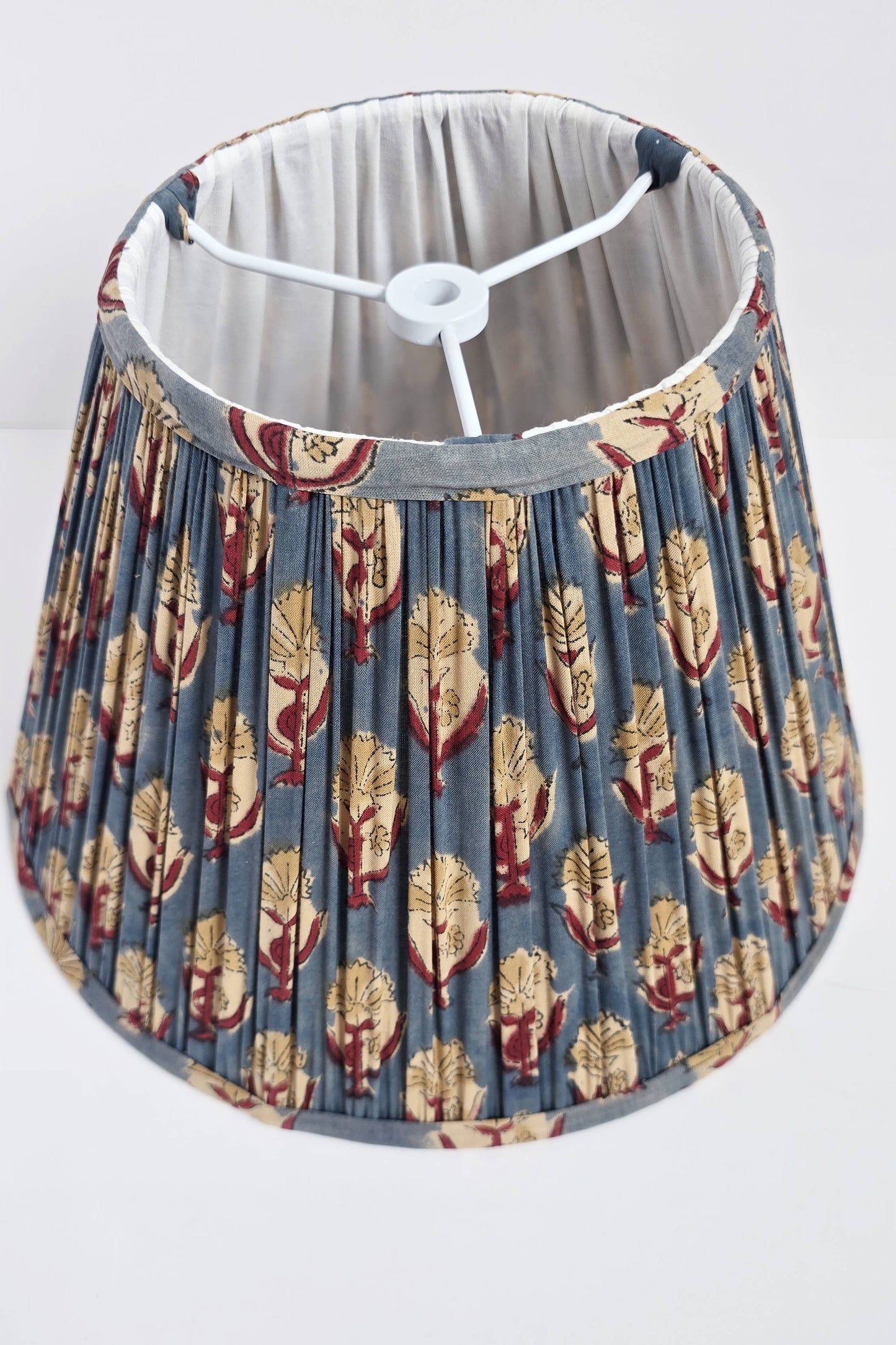 A pleated empire lampshade with blue and red floral blockprint patterns, displayed on a table next to a framed picture and a decorative object.