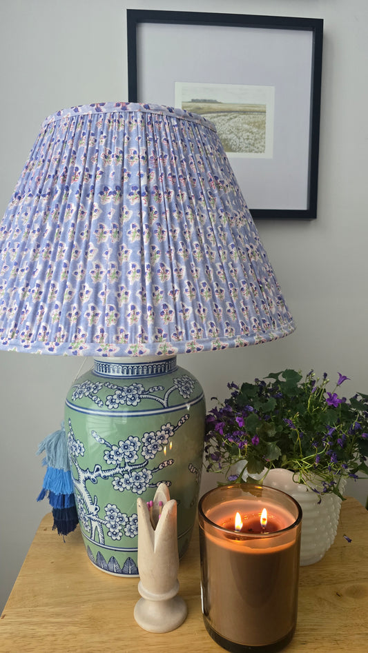 Decorative lamp with a patterned shade on a table with candles and a plant.