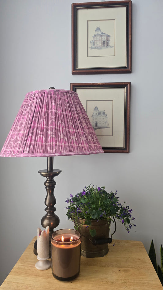 Lamp with pink shade, candle, and plant on a wooden surface with framed pictures on the wall.