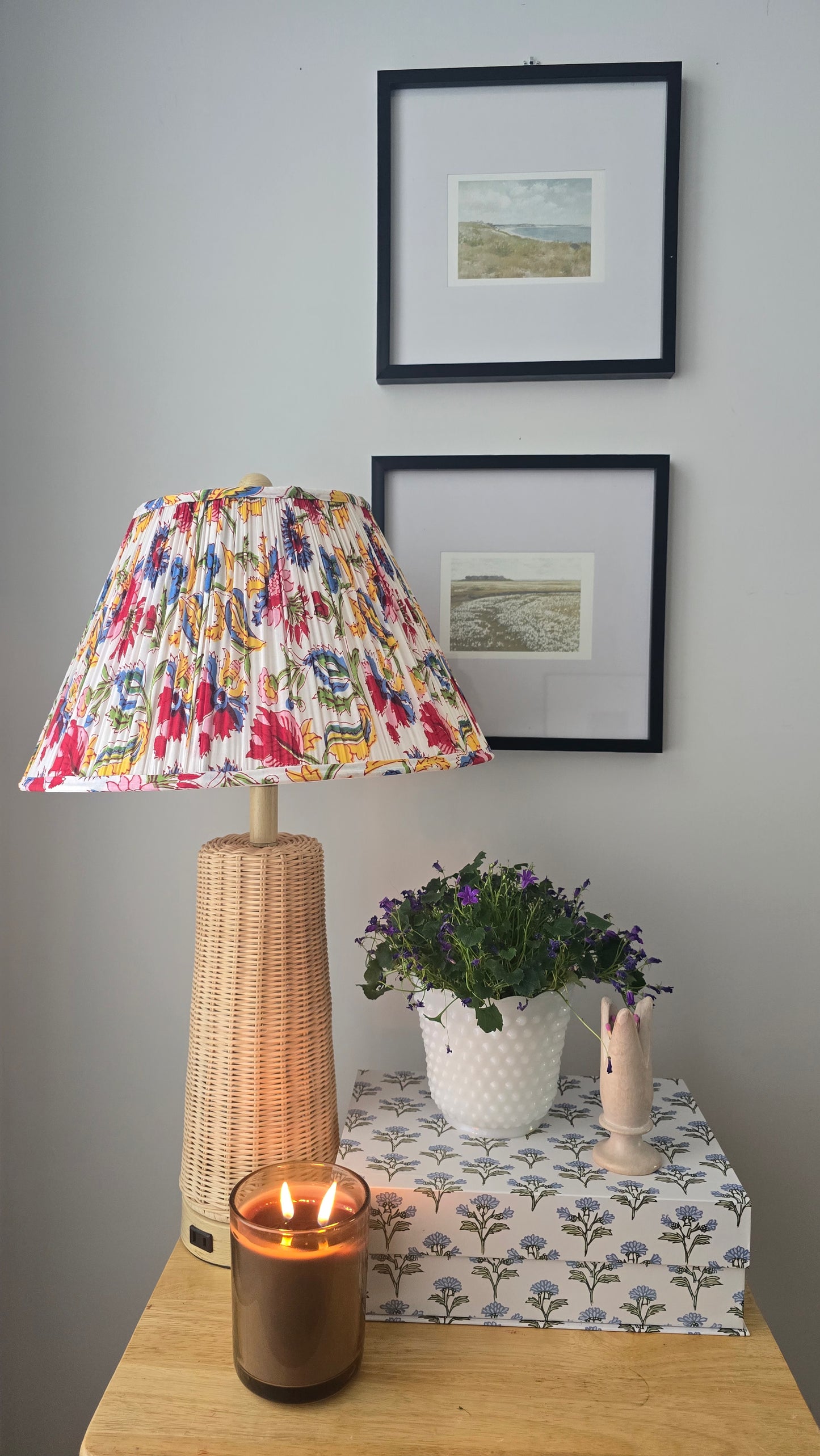Decorative lamp with a colorful shade on a table next to a candle and a plant, with framed pictures on the wall.