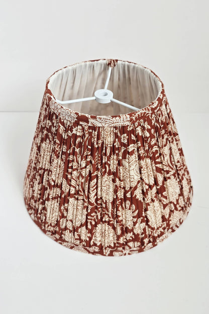 A pleated empire lampshade with a brown floral blockprint pattern, displayed on a table next to a framed picture and a decorative object.